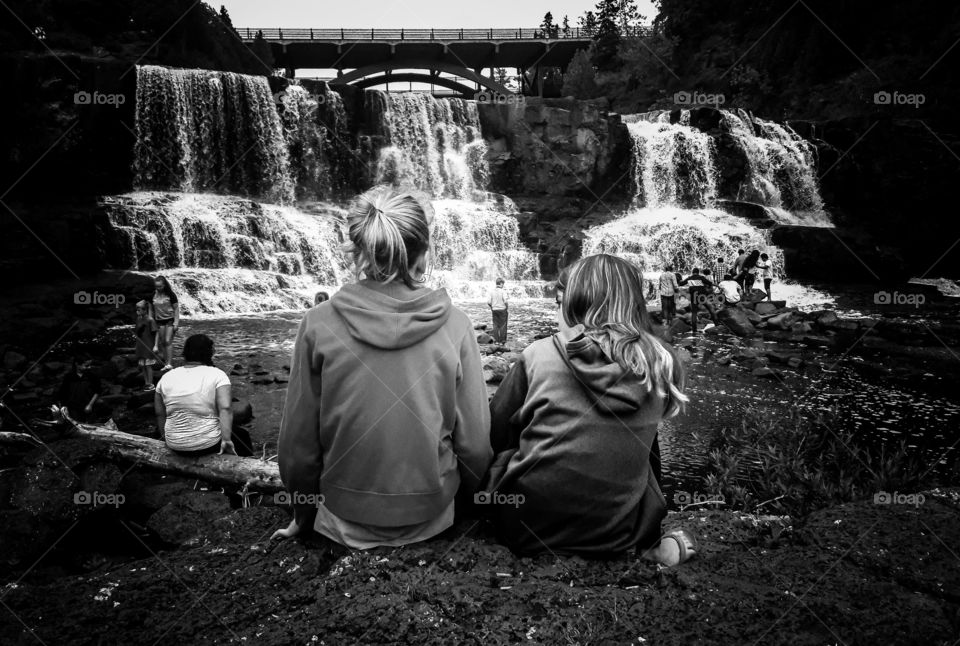 Black and white photograph of my daughter and, her friend. Sitting in front of a waterfall at Gooseberry Falls, located in Minnesota.