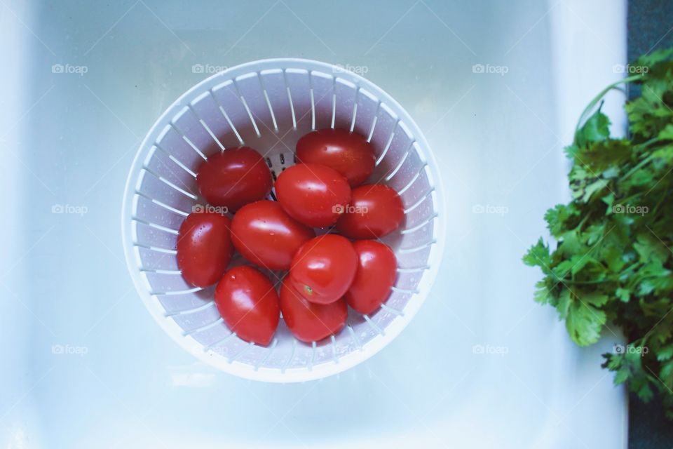 Roma tomatoes in a white colander in a white sink, freshly picked cilantro to the side on a green countertop