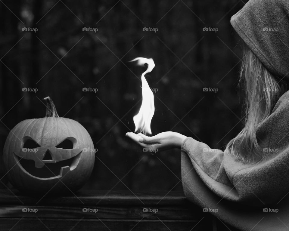 Black and White; Cloaked woman holding a large (real and safe) flame in her hand that’s pointing to a carved pumpkin