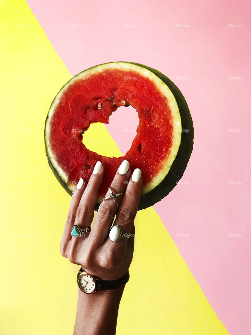 Female hand with white nails and fashionable rings and watches holding a slice of watermelon on colorful pastel background 