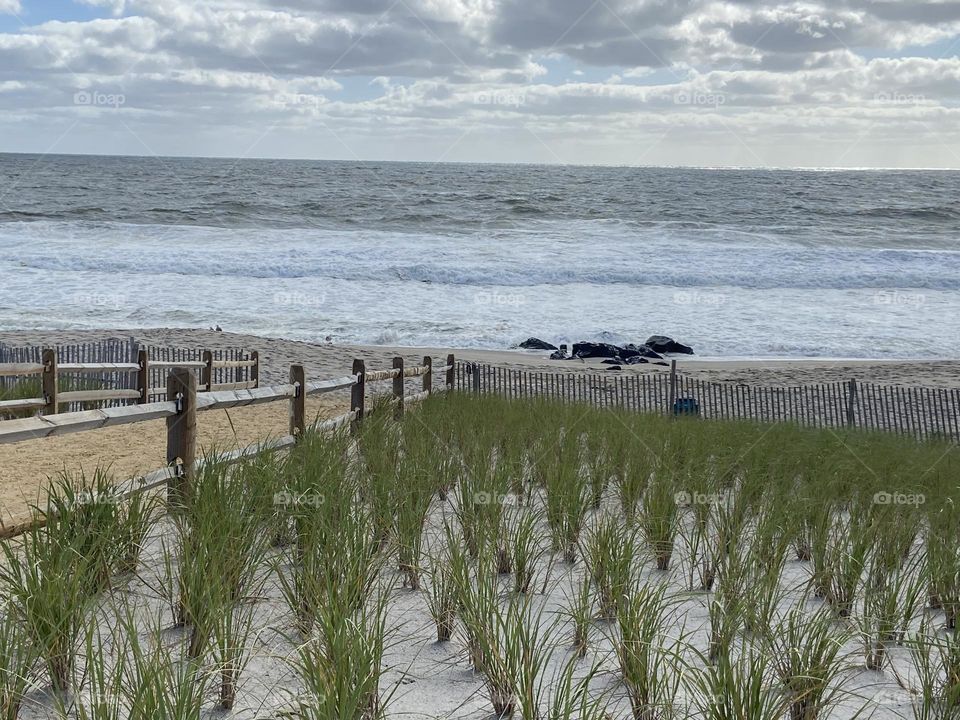 A view of the ocean and beach in Bay Head, NJ from an elevated platform with benches that leads to a sand path to the beach. Dune grass adds some green to the scene. Clouds fill the sky above the ocean on this September day. 