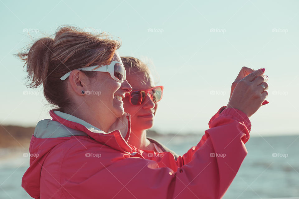 Young woman taking a photos using phone, looking at screen, standing outdoors, she is backlighted by sunlight with plain sky and sea in the background