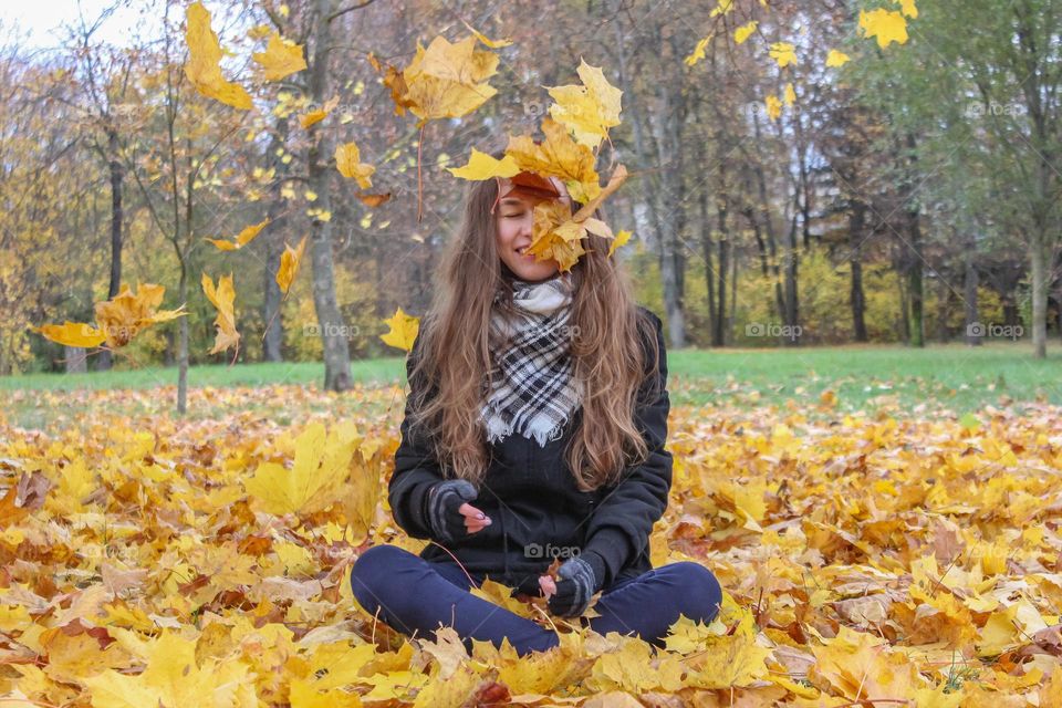 The girl is sitting on the autumn yellow leaves and leaves are falling on her from above.