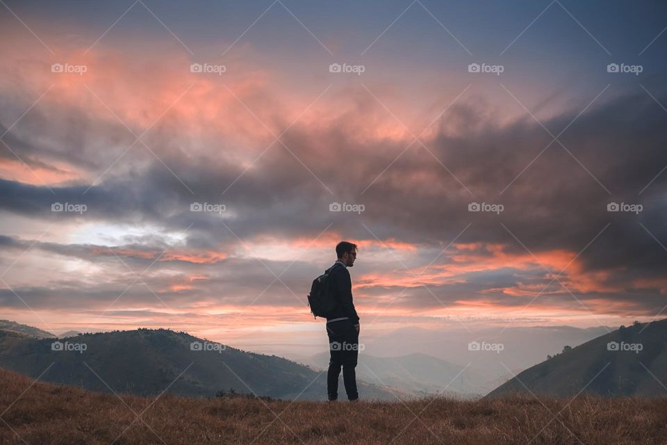 Rear view of male hiker standing on field against sky during sunset. 
