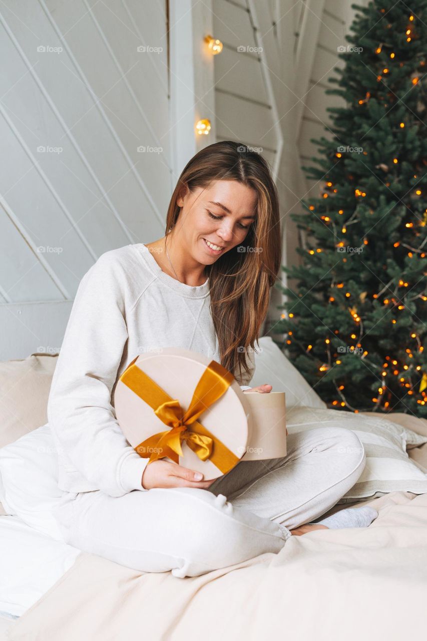 Young beautiful woman with dark long hair in cozy white pajamas with present gift box withgold ribbon sitting on bed in room with Christmas tree at home, Happy New Year