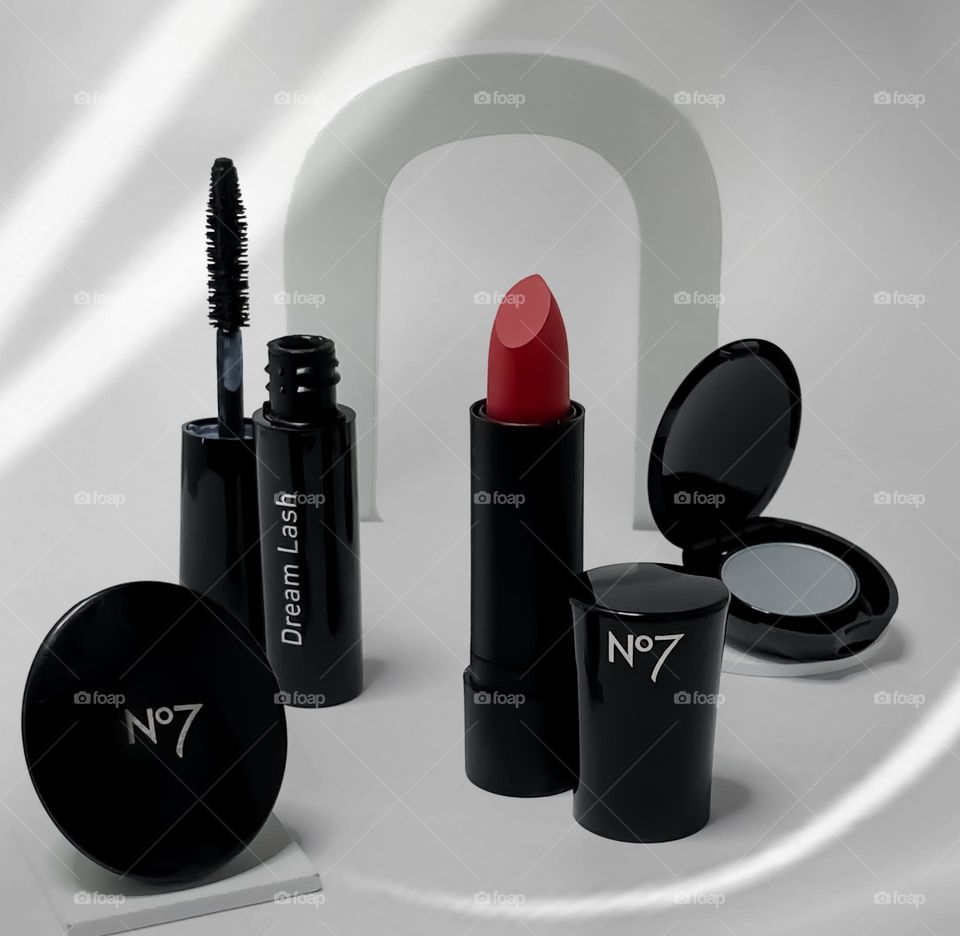 A collection of Boots No7, black cased cosmetics against a white background with an arch over red lipstick and subtle shadowy overlay