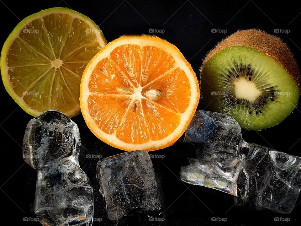 Fruits cut in half: green kiwi, orange orange and yellow lemon, which lie among ice cubes on a black background