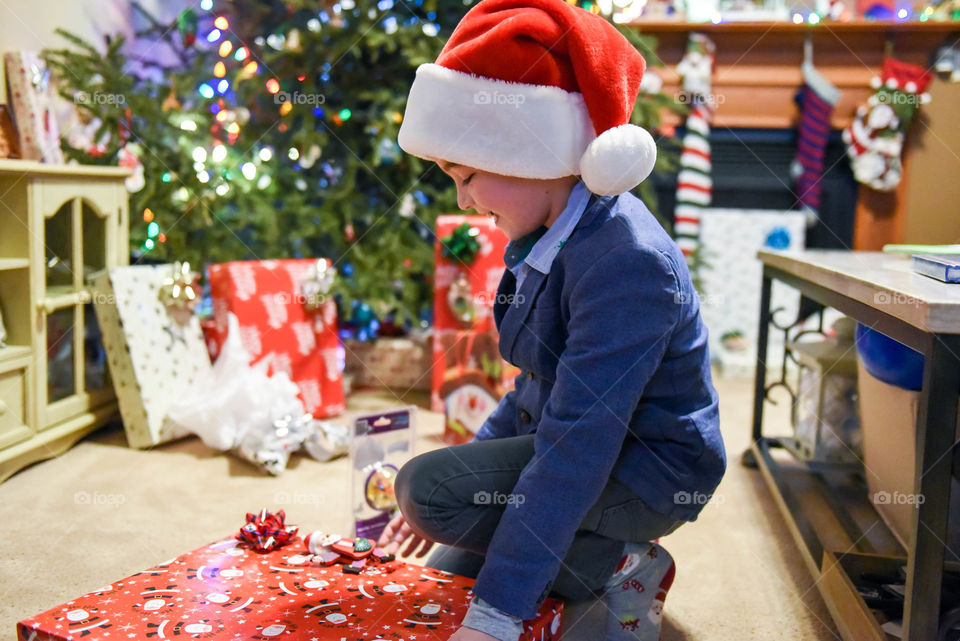 Young boy wearing a Santa hat and opening a Christmas present in a decorated home