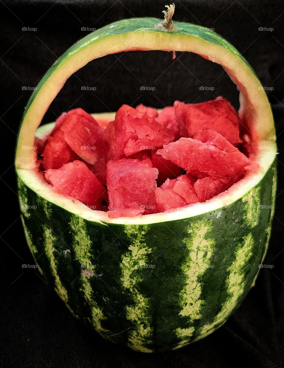 Summer treats. A green watermelon rind with black stripes is cut into the shape of a vase with a handle. Inside are juicy slices of red watermelon. Black background