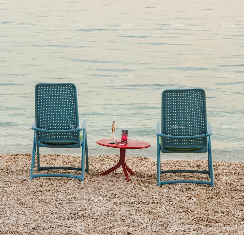 Two blue chairs with a red table in the middle standing on the pebble beach of the Adriatic Sea in summer on a warm, windless evening