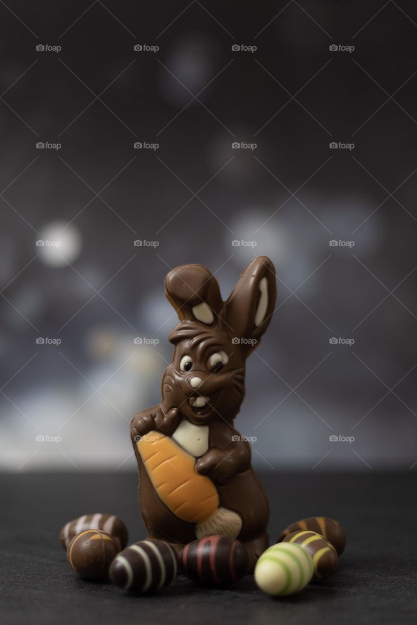 One cute and happy chocolate bunny with an orange carrot in its paws and candy eggs around it stands on a black stone table, close-up side view. The concept of a holiday treat,happy easter.