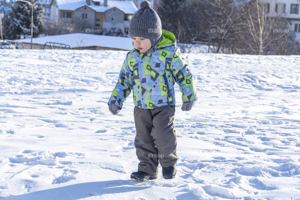 A child with a serious expression on his face in winter clothes jackets, pants, hat and boots in winter on the white snow on the street and in the park in nature plays winter fun.