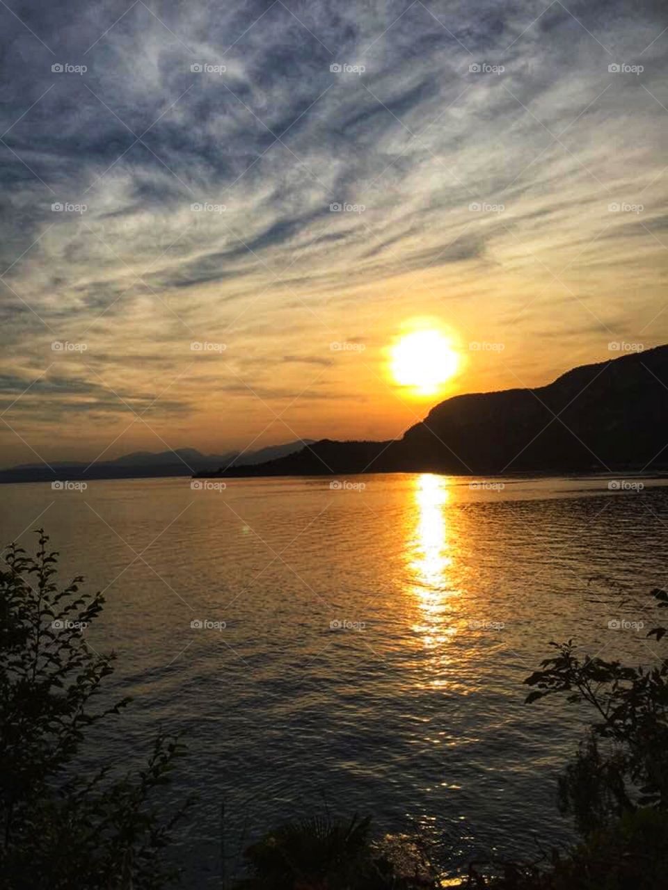 Beautiful sunset on the lake . The sun reflected into the water with a long orange streak . The trees and mountains turning dark. sky there are many gray clouds