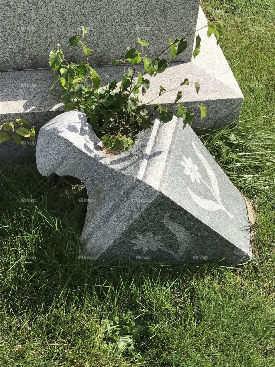 The decorative top of a monument has broken off and is laying in the grass.