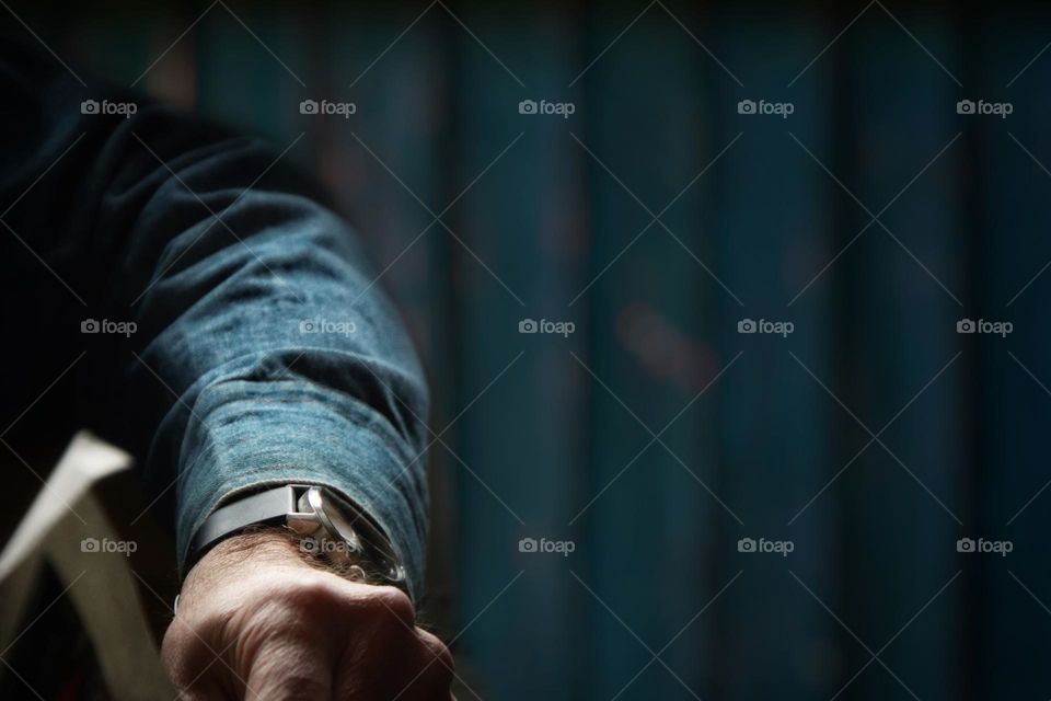 An arm with a wristwatch in a blue denim jacket and a hand holding a newspaper in front of a blue wooden wall