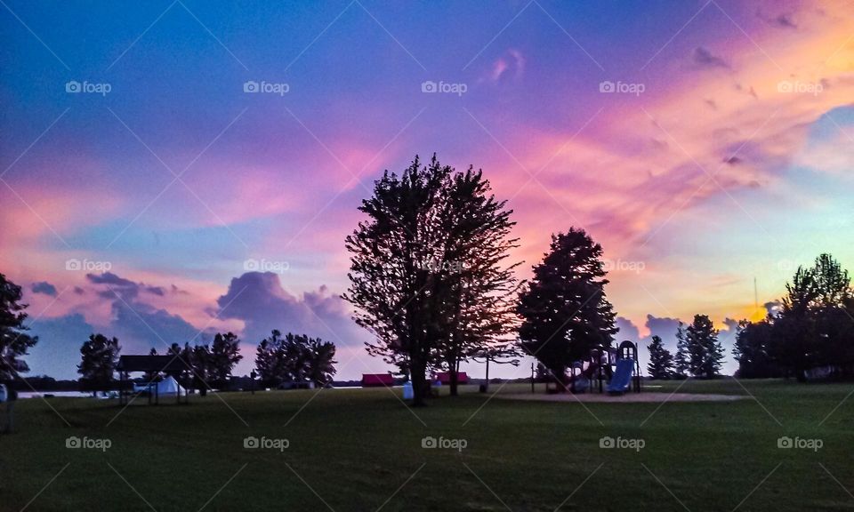 Sunset sky in brilliant purple, pink, orange, yellow, and blue making the silhouette of the trees pop in a very green open field. 