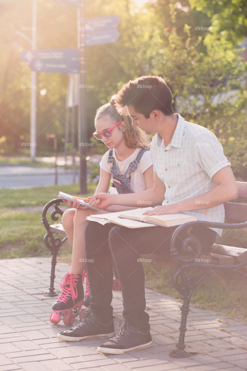 Students making the notes learning from books sitting on a bench in a park. Young boy wearing a blue shirt and dark jeans. Young blondie girl wearing jeans and sunglasses