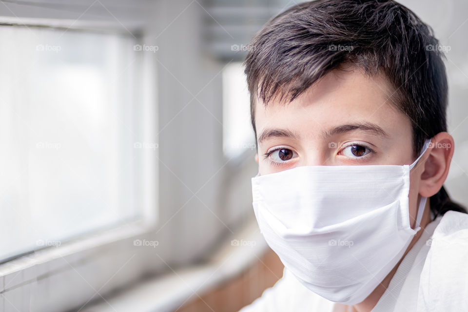 Child in a white protective mask near the window. House pandemic period. Concept of the fight against coronavirus COVID-19.