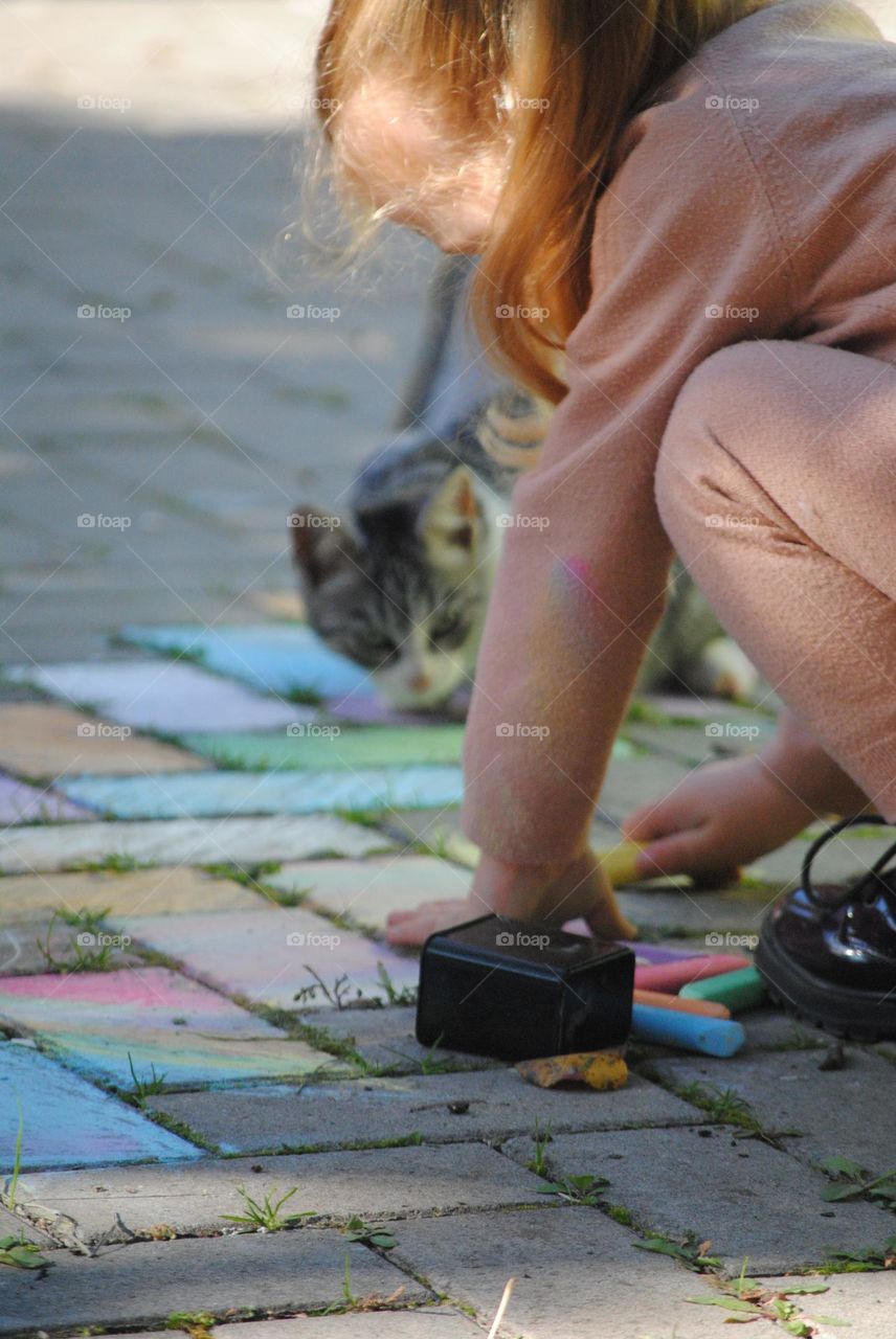 This picture represents a little girl drawing with chalk on the asphalt, and a cute kitten that is very attentive to what the little girl is doing