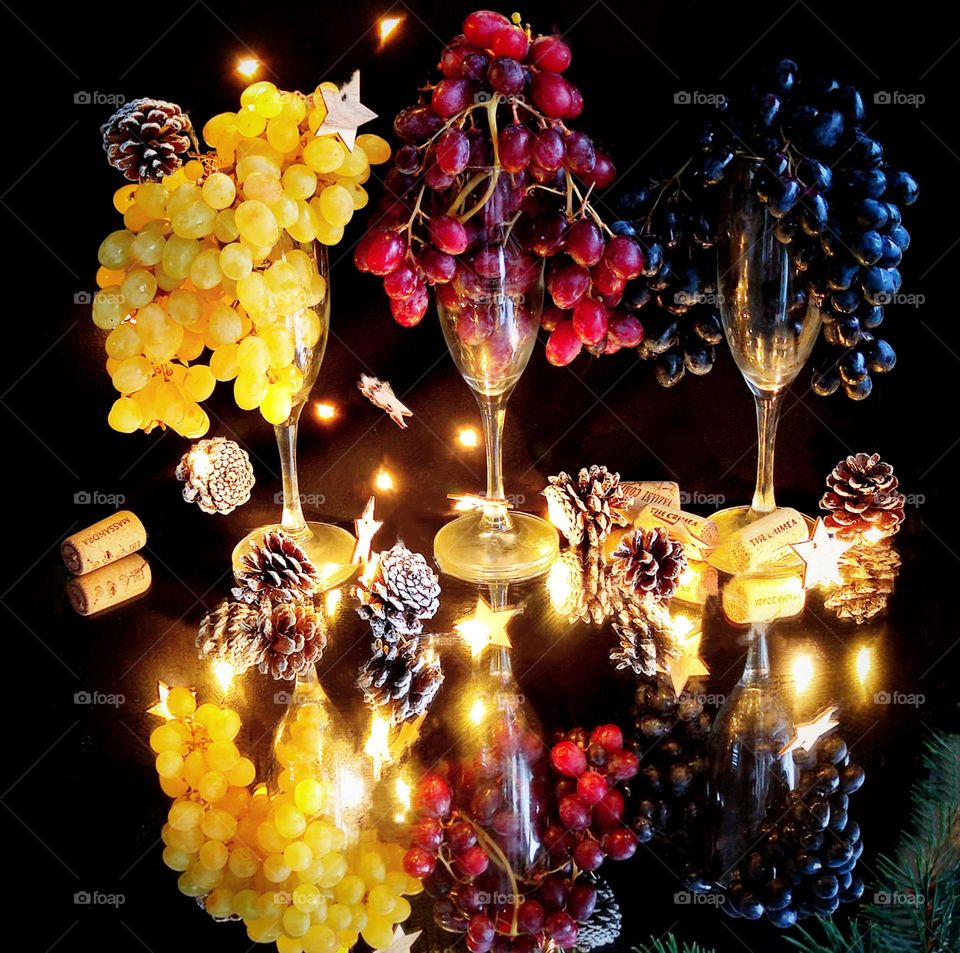 Three glasses with bunches of grapes in the form of hair stand on a mirrored surface.  Yellow, red and black grapes.  Around a luminous garland, pine cones and stars.  Wine corks lie at the stems of the glasses.  Reflection in the mirror