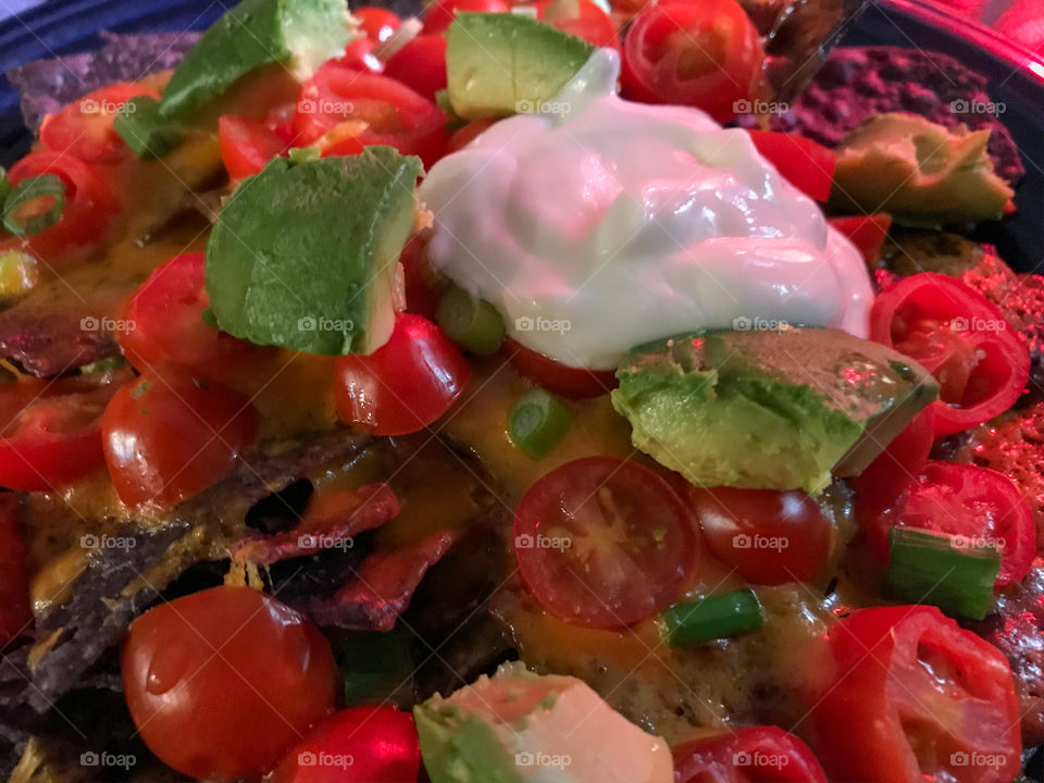 Who doesn’t like Nachos?! Closeup of blue corn chips, smothered with melted cheese, sliced grape tomatoes, green onions, chunks of ripe avocado, a dollop of sour cream & a splash of hot sauce! Delicioso!