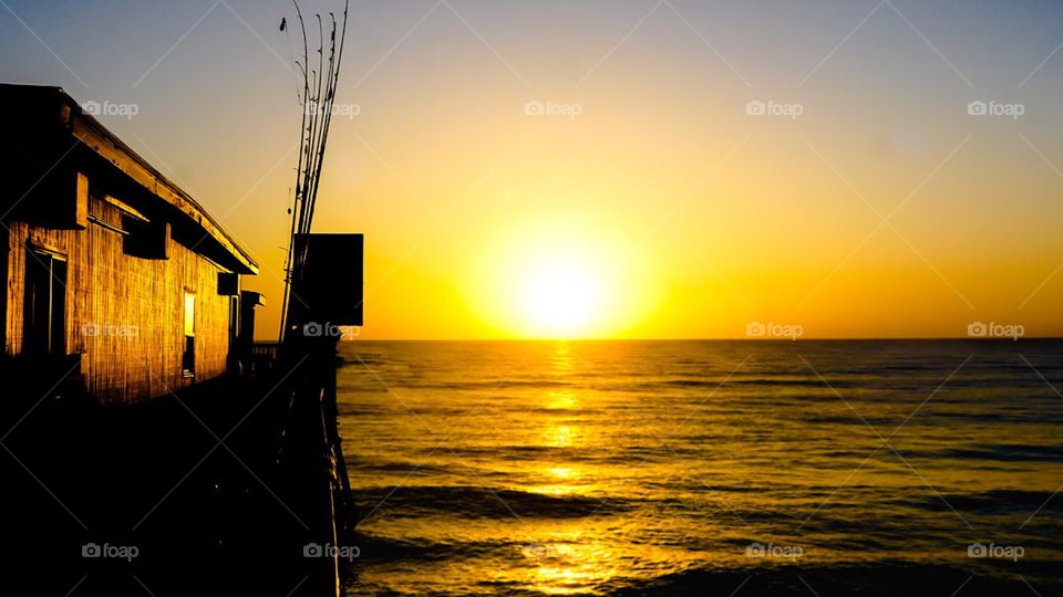 Fishing poles on the fishing pier are silhouetted by the brilliant yellow morning sunrise over the calm Atlantic Ocean. 