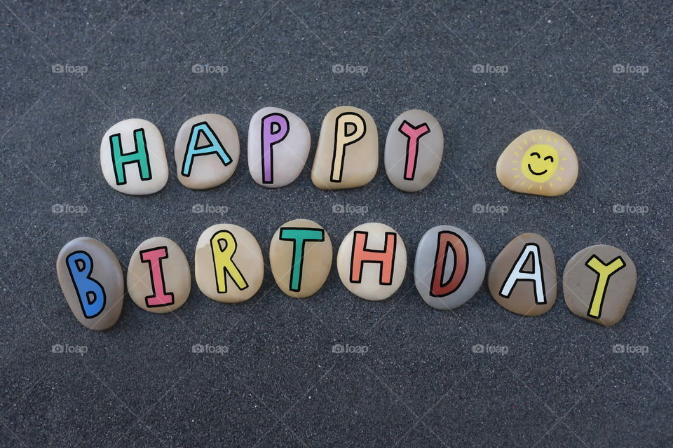 Creative happy birthday with colored letters on sea pebbles over black volcanic sand