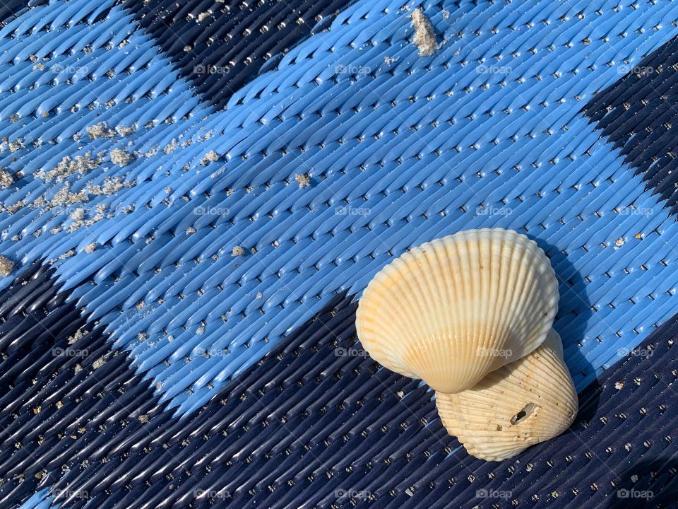 Two seashells on a blue sand proof carpet with geometric shapes dark and light blue colors with some sand representing the leisure and enjoyment of time spent at the beach on vacation.