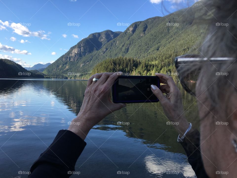 Middle aged woman in sunglasses taking photo if beautiful Canadian mountain lake scene with smartphone