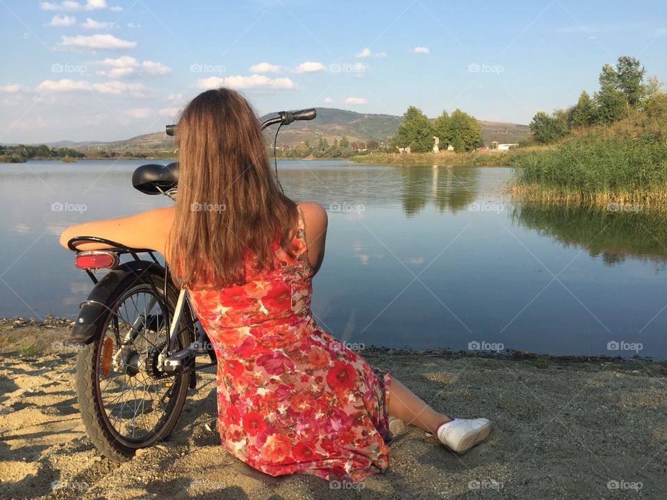 Back of woman wearing flower dress sitting on the sand looking at the lake with bicycle near her