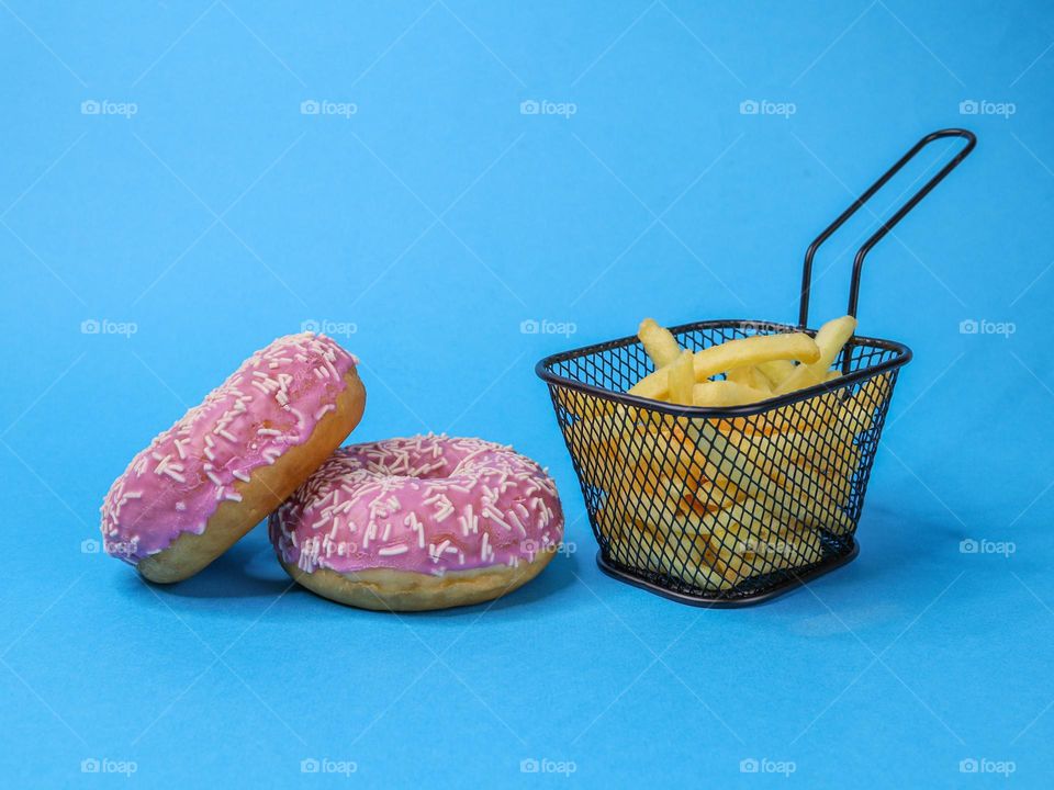 Two American donuts with pink glaze and French fries in an iron black basket lie on a wall, close-up side view. Fast food concept, unhealthy food.