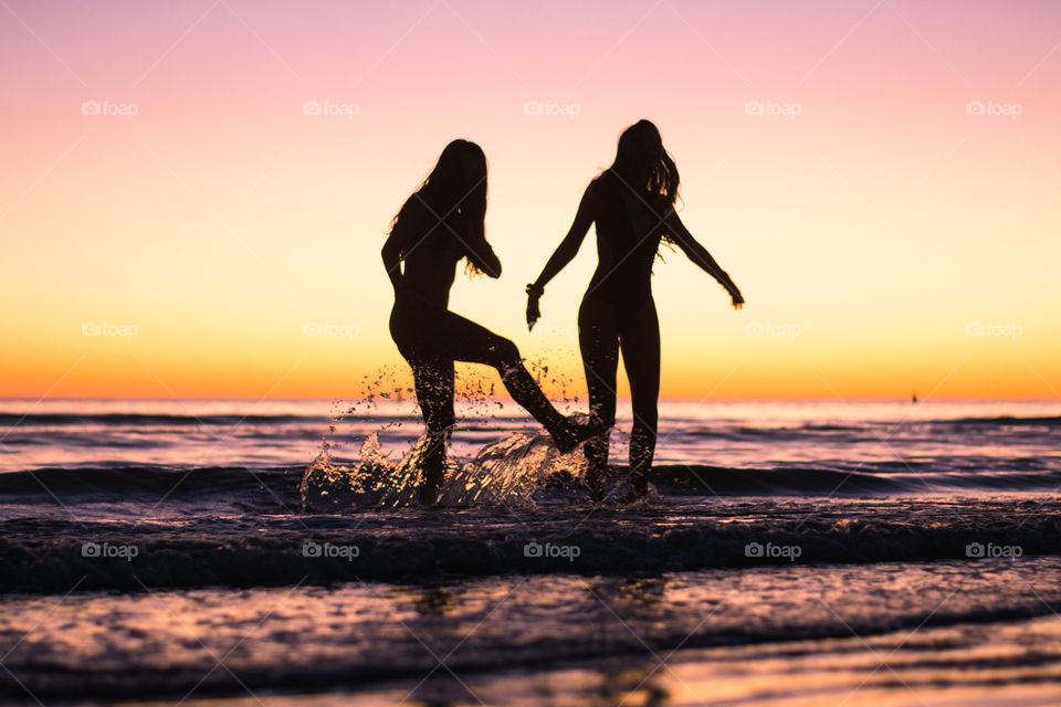 Two girls play and dance in the ocean by the beach during sunset with beautiful pink skies around them
