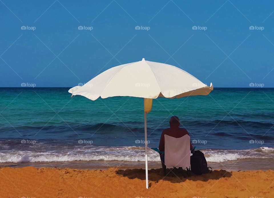 man on a beach under umbrella near brilliant blue sea and honey coloured sand