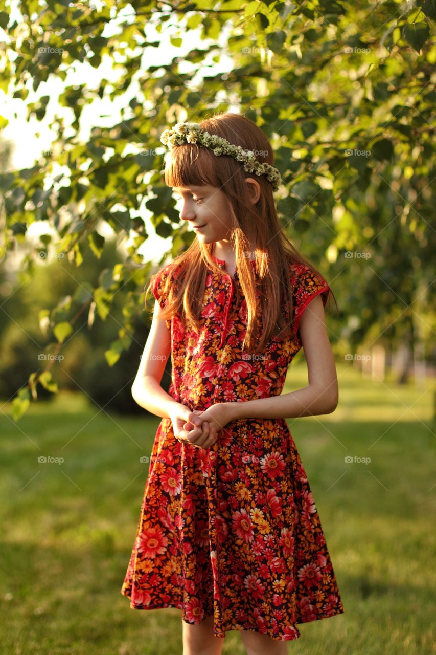 Little girl in a red dress with a wreath of clover on her head with long and blond hair smiles in the sun in a park full of green foliage