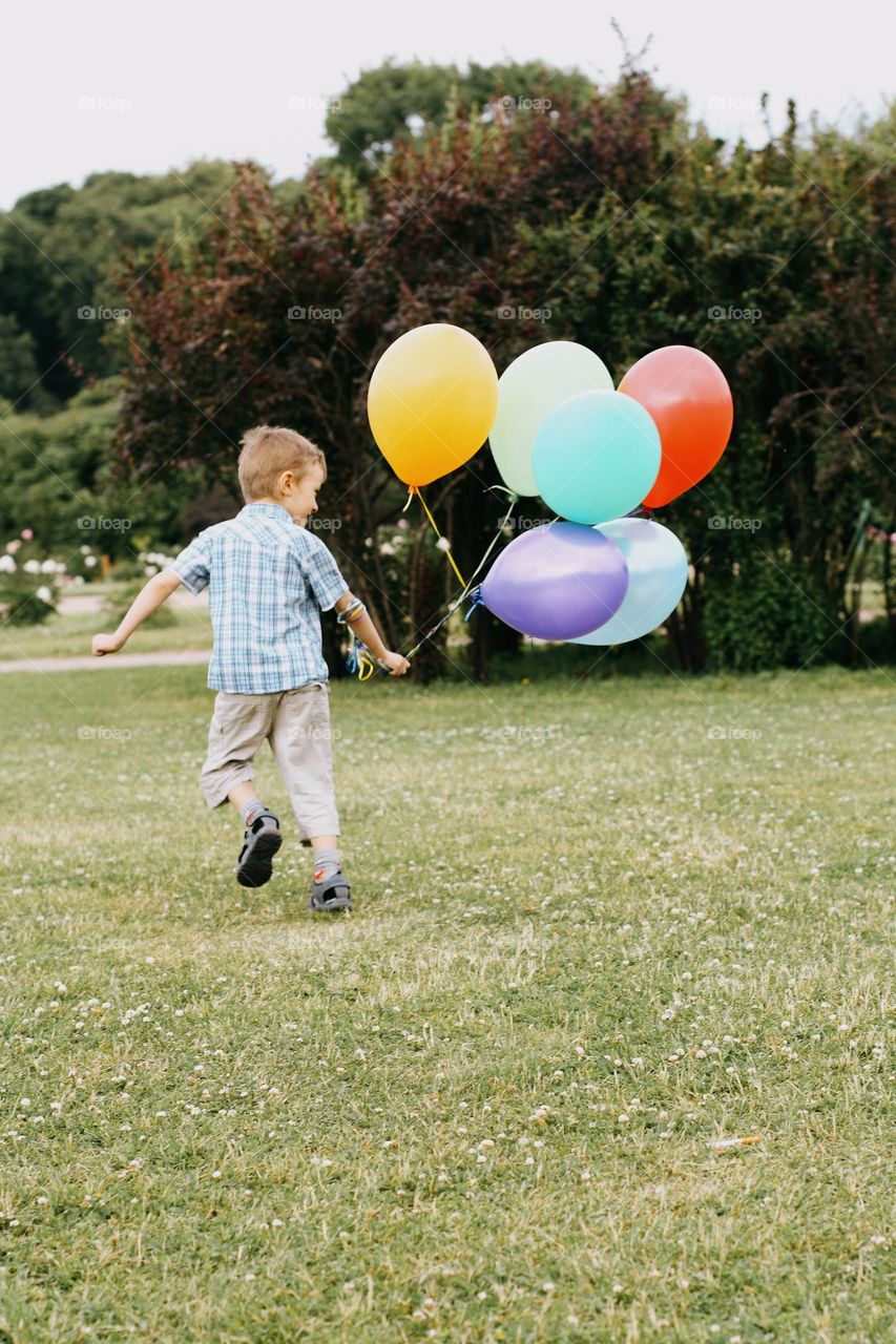 A little blond boy in a white T-shirt running on a green field, holding a bunch of colorful balloons in his hand, back view, LGBT colors