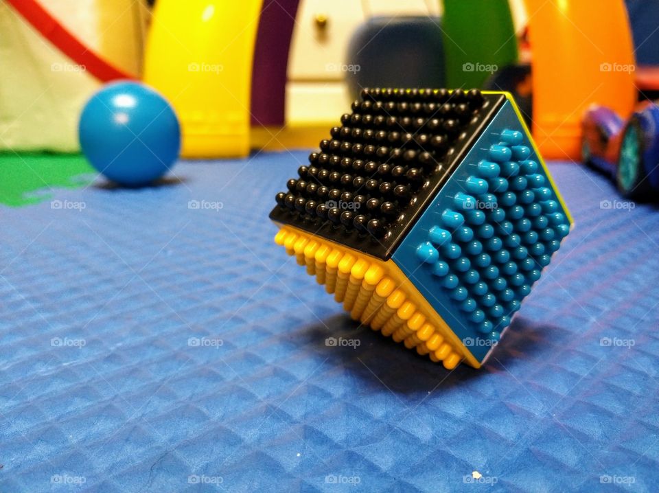 A colorful building cube resting on one edge in a child's play room, with a blue ball in the background.