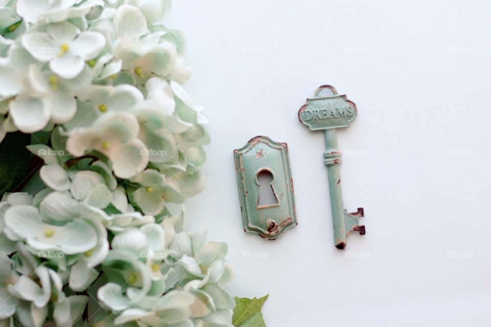 Overhead view of light teal, vintage, metal key with embossed word, “Dreams,” and keyhole with light teal hydrangeas on white background -