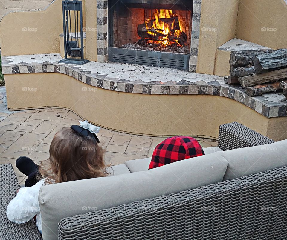 A view from behind shows two young children sitting on a couch enjoying the warmth of a backyard living space fire. No faces are shown therefore the children not recognizable.