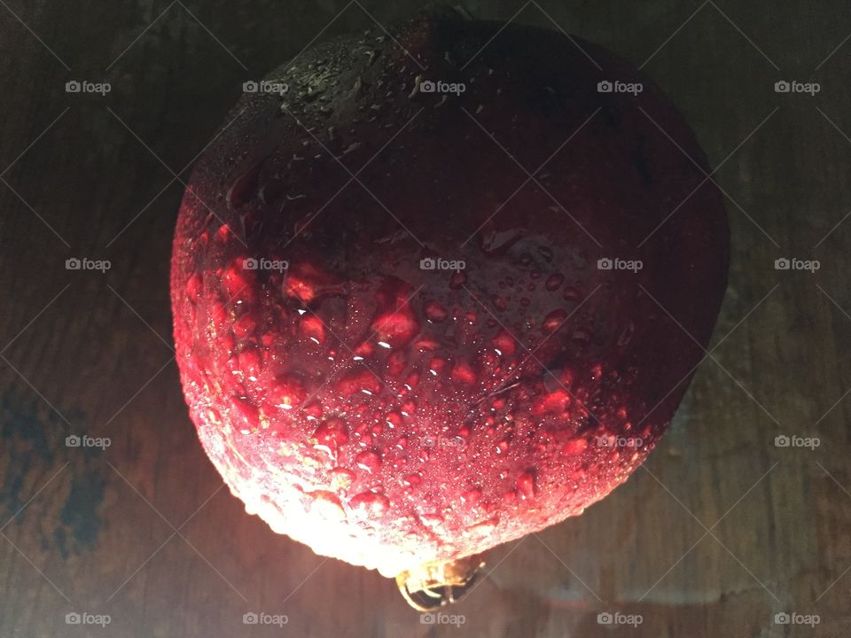 Still life of a wet whole pomegranate lit from the side. The water drops glow red on the red skin. Looks like a planet orbiting past its sun. 