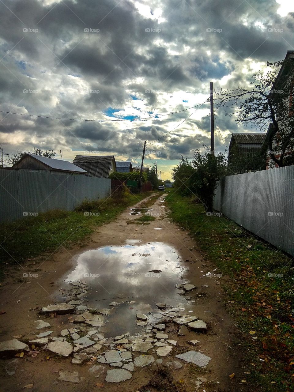 View of the road in the village and a puddle on this road, which reflects the cloudy sky, on a cloudy autumn evening