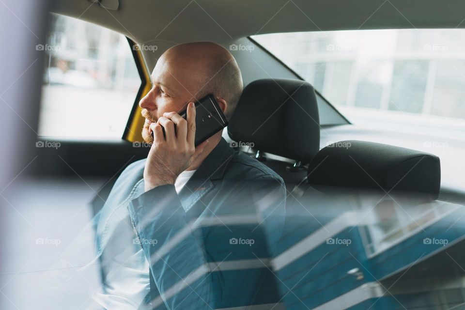 Handsome bald bearded man businessman in suit using mobile phone inside yellow car taxi at city