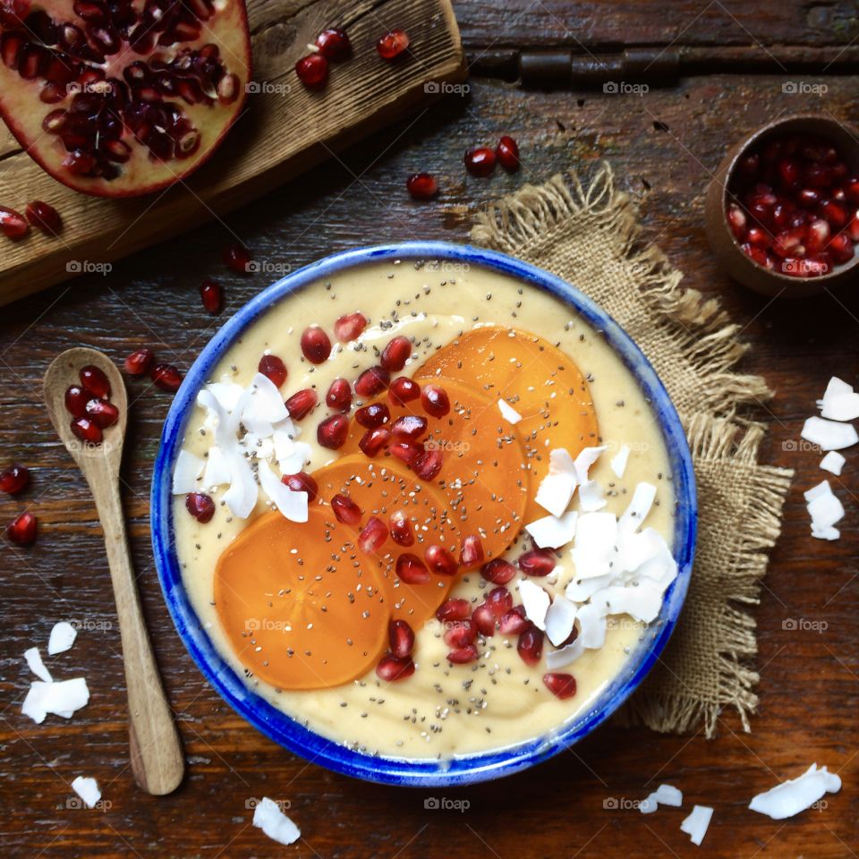 Banana and persimmon smoothie bowl with persimmon slices, pomegranate and coconut flakes in a blue bowl in a rustic wood table.