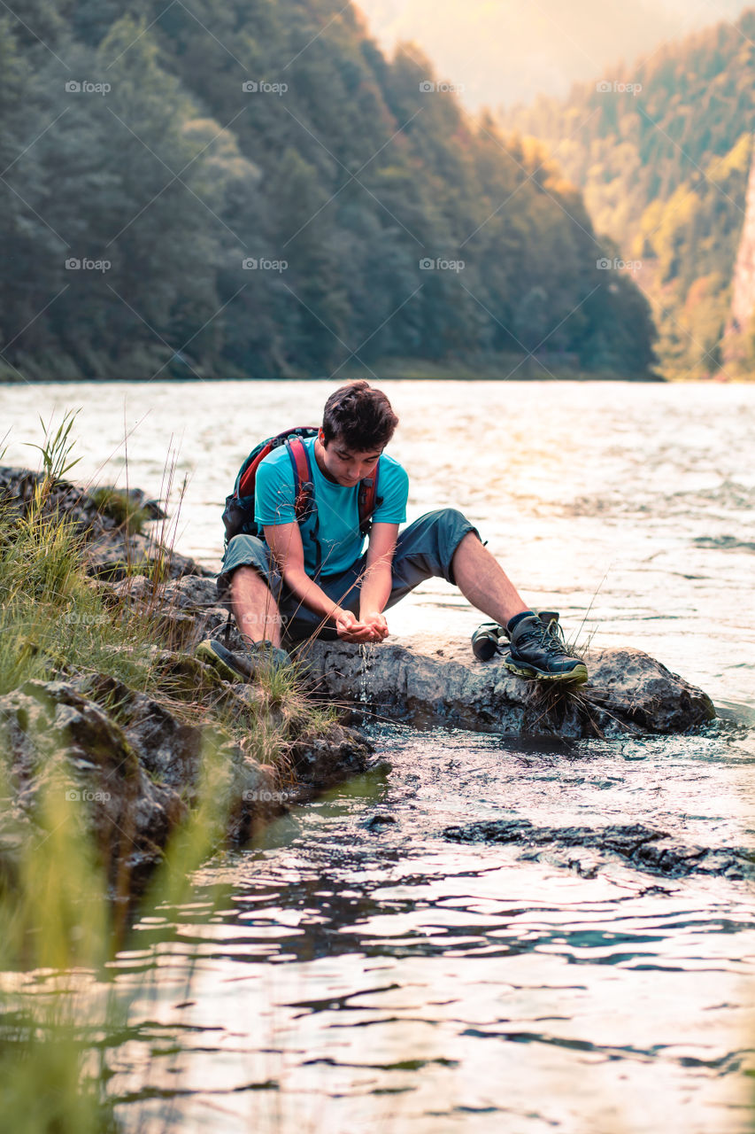 Young boy takes pure water from a river and holds it in the hands. He is sitting on a rock over the river, rests during a hike, spends a vacation on wandering with backpack, he is wearing sport summer clothes