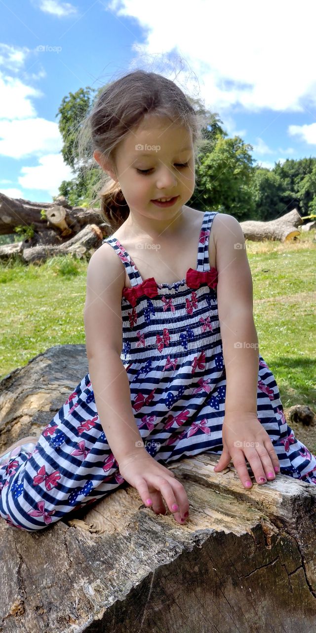 little girl in dress sat on log in park in bright sunshine