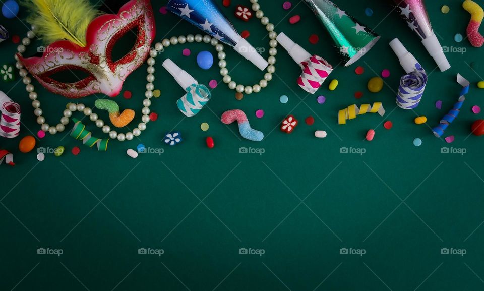 A masquerade mask, a white pearl necklace, whistles, pipes, jelly candies and colorful confetti lie on top of a dark green background with copy space below, flat lay close-up. Concept for masquerade, festival, carnival, party banner.