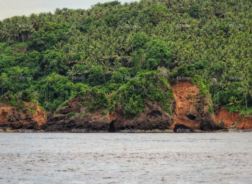 Natural scenery, three large rocks lined the beach, large rocks with grass and green plants. Background of a hill with green trees.