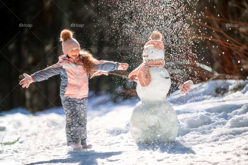Little girl with snowman in winter forest at sunny day