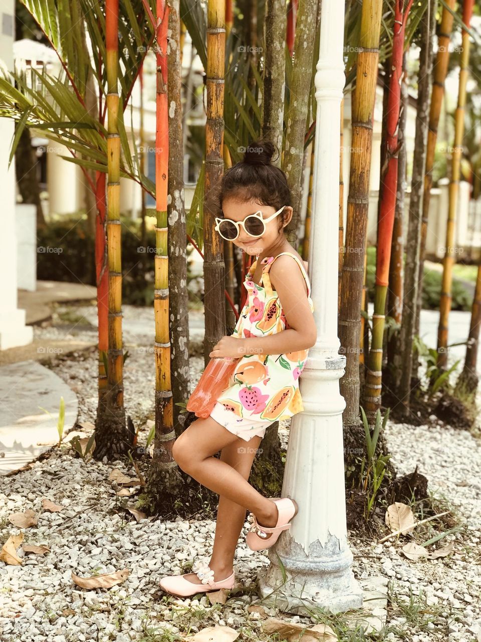 A cute smiling girl lean over a pole , looking towards a down and smiling and she is in summer outfits and holds health drink .