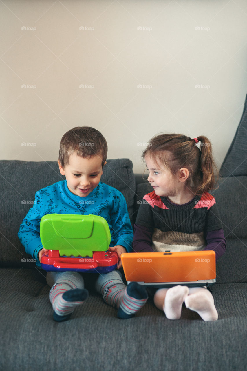 Two children playing with laptops learning basic digits, characters, sounds and images. Little girl and boy sitting on sofa and playing together at home. Candid people, real moments, authentic situations