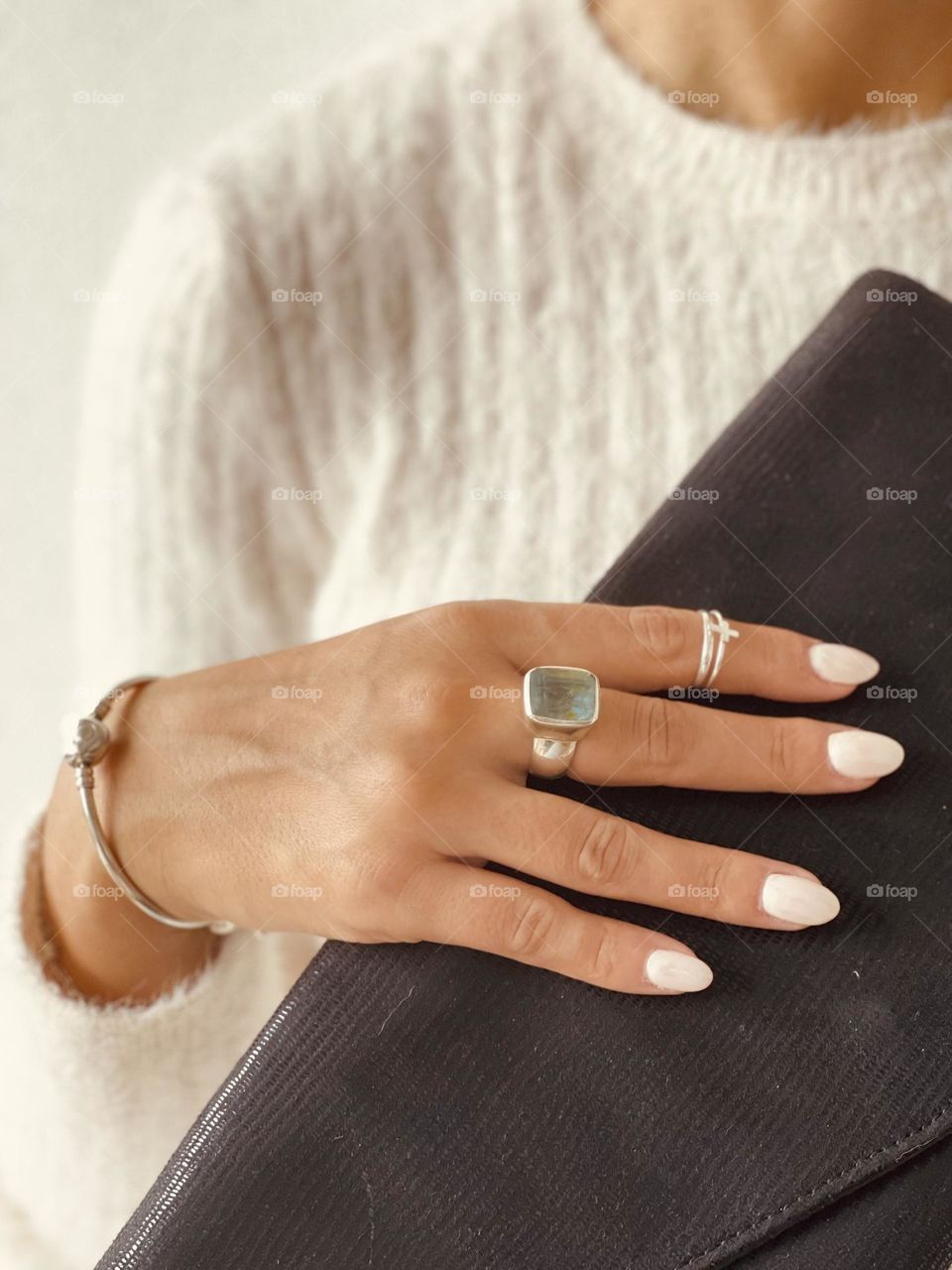 Woman's hand wearing white nail polish, silver bracelets and rings with aquamarine stone while grabbing a black clutch bag